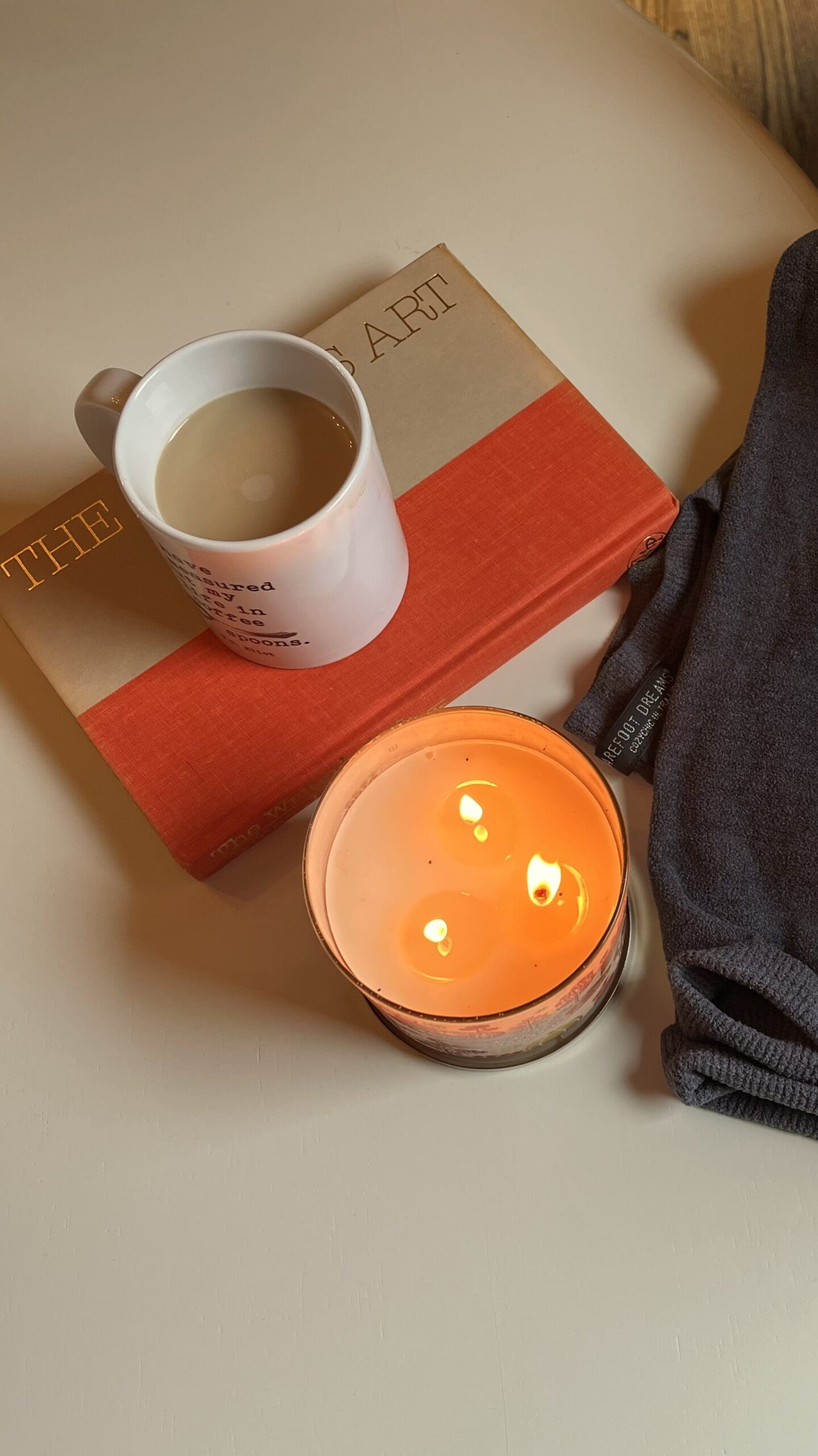A Barefoot dreams sweater next to a book, a cup of coffee, and a candle