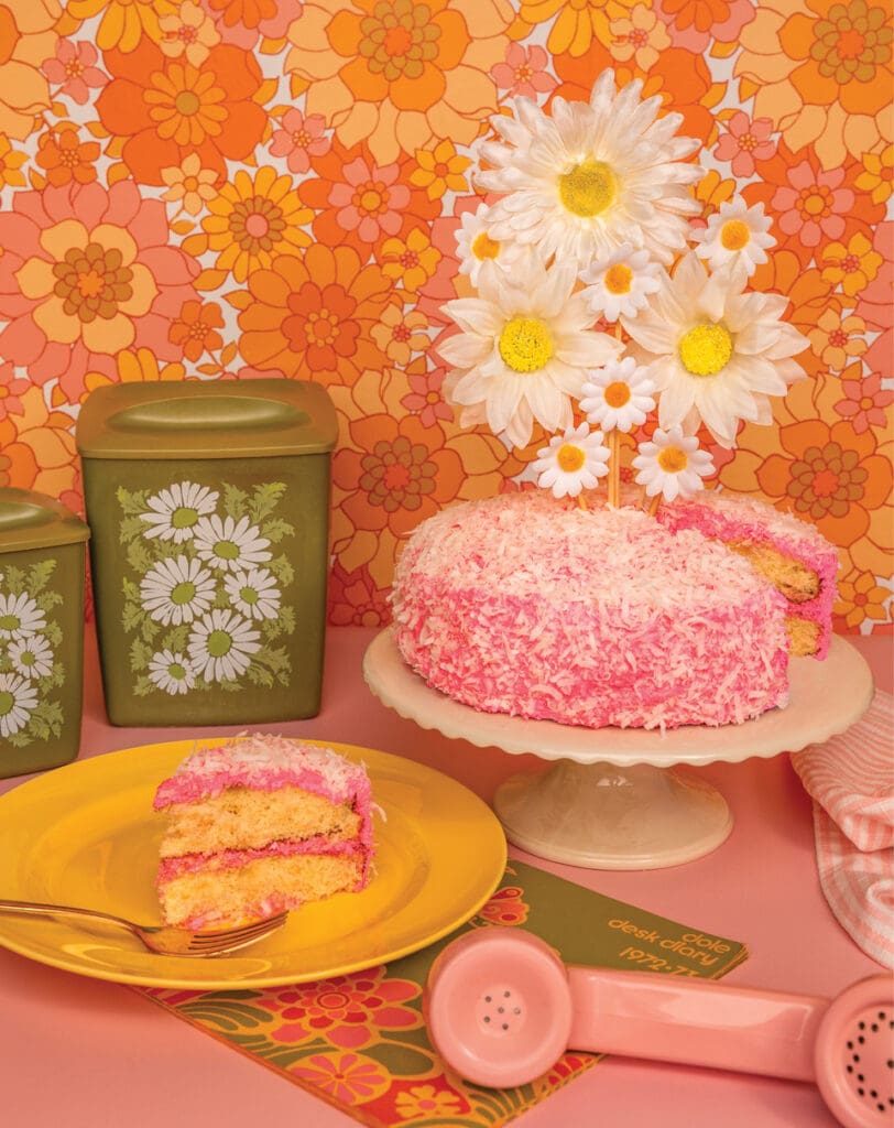 Pink coconut layer cake frosted in bright pink icing and covered in shredded coconut, styled in a retro 1950s kitchen setting.